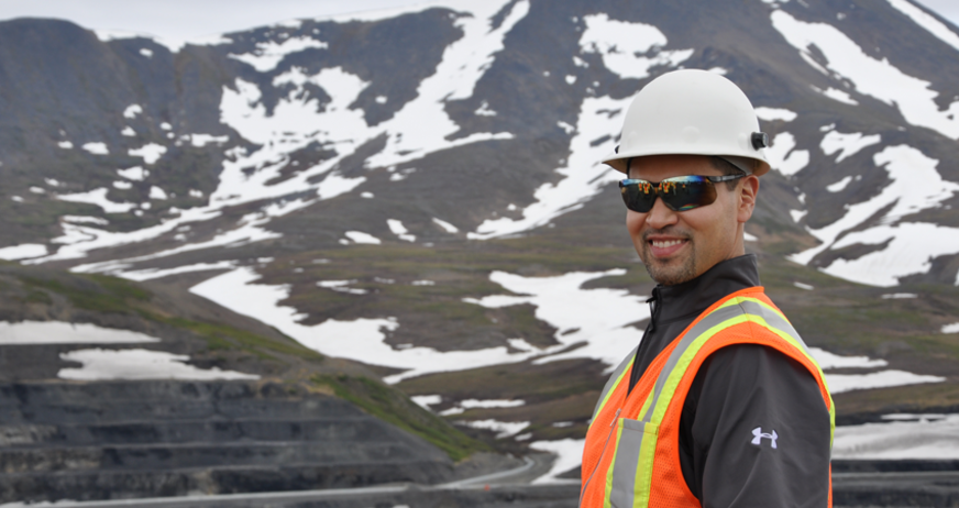 Smiling worker in safety gear stands in front of snowy, mountainous landscape at an industrial site.